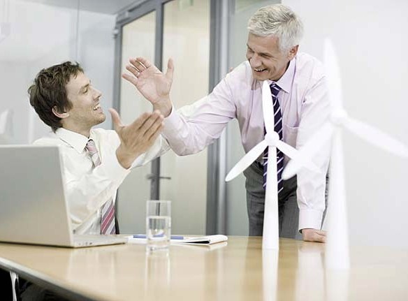 Two businessmen sitting in an office with model wind turbines celebrating success